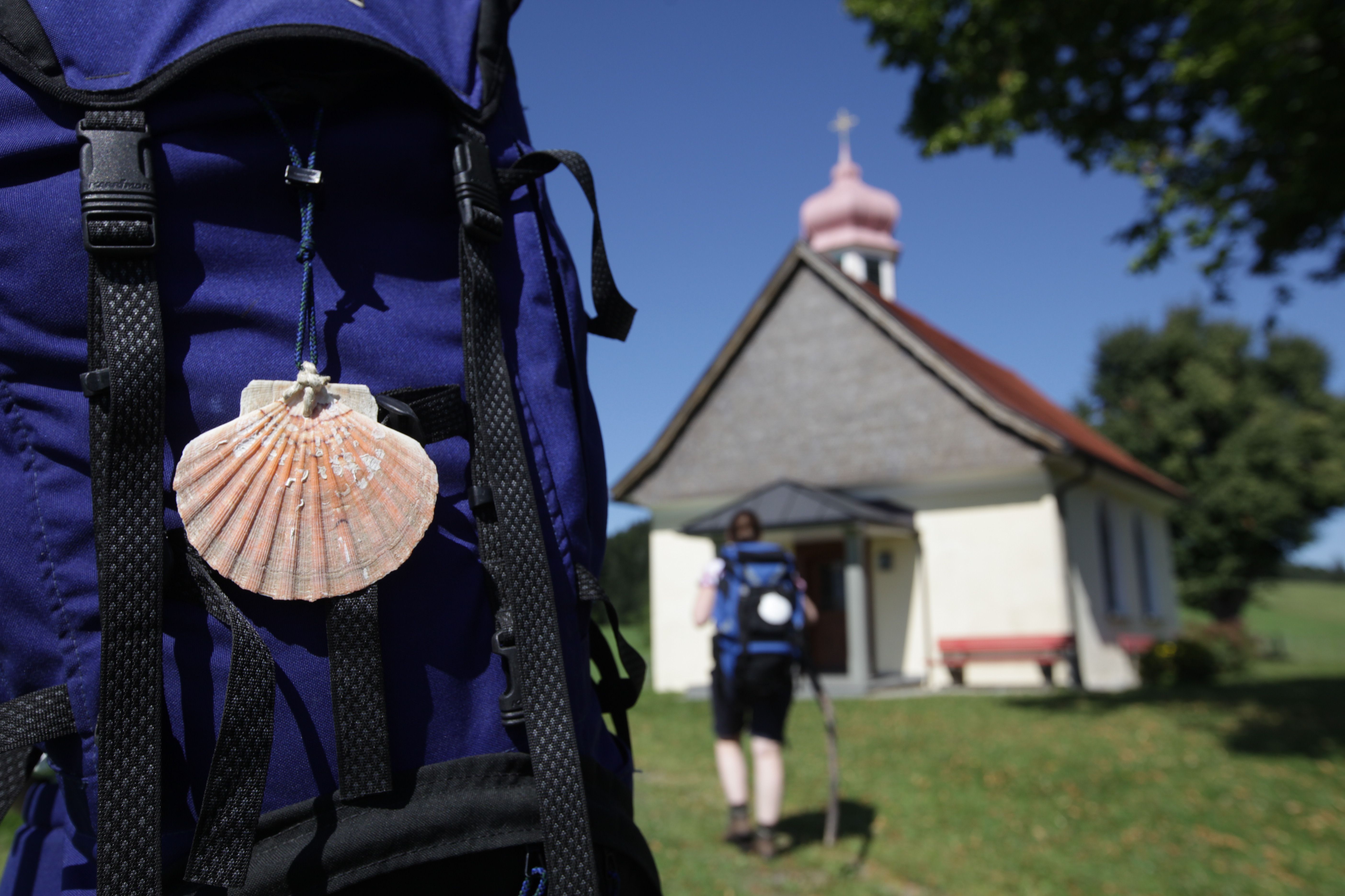  Im Vordergrund steht ein Rucksack mit einer Muschel als Anhänger und im Hintergrund ist eine Kapelle zu sehen - das Bild wird mit Klick vergrößert 