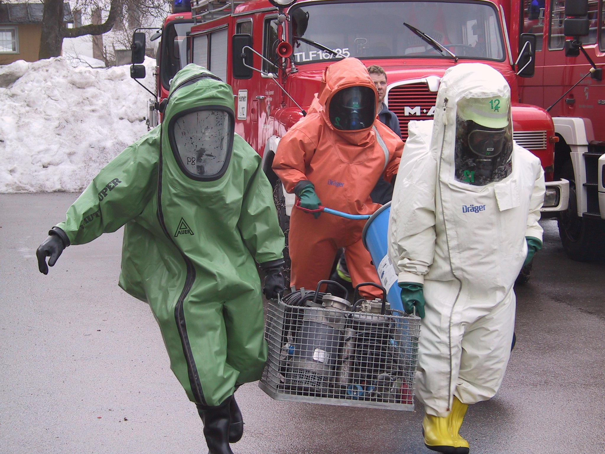  Drei Personen tragen jeweils einen grünen, roten und weißen Ganzkörperschutzanzug und transportieren einen Korb. Im Hintergrund ist ein Feuerwehrauto zu sehen - das Bild wird mit Klick vergrößert 