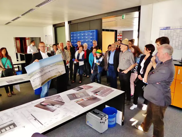 Mehrere Leute stehen in einem Büro der Holzer Druck und Medien Druckerei. In der Mitte steht ein Mann und zeigt ein Plakat in die Runde (Das Bild wird durch klicken vergrößert)